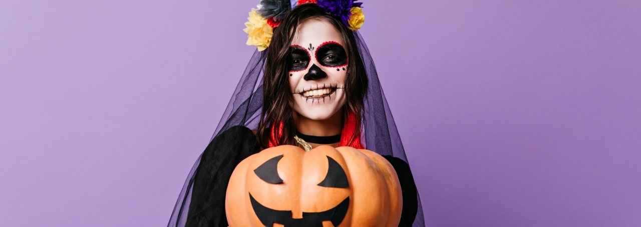 Imagem de mulher com rosto maquiado para o dia dos mortos, segurando uma abóbora, símbolo do Halloween