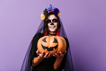 Imagem de mulher com rosto maquiado para o dia dos mortos, segurando uma abóbora, símbolo do Halloween